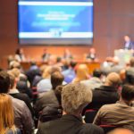 image of a conference audience and speaker to show Why Your Closing Question Fails Moving from Hero Performers to Scalable Systems